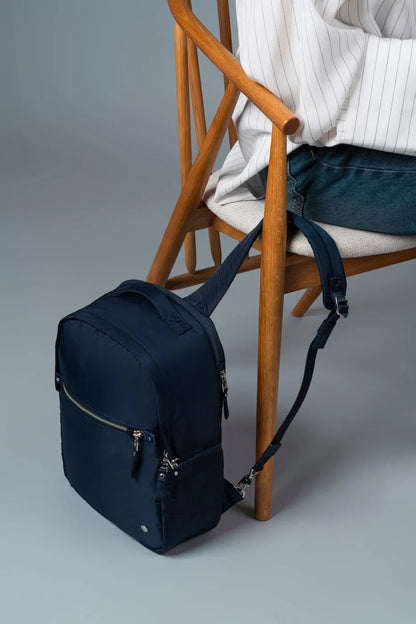 Pacsafe W 10L Anti-theft 13" Laptop Backpack in Navy, tethered to a chair leg by its strap while its owner sits