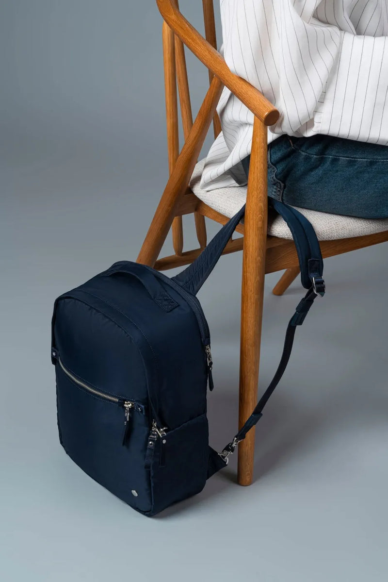 Pacsafe W 10L Anti-theft 13" Laptop Backpack in Navy, tethered to a chair leg by its strap while its owner sits