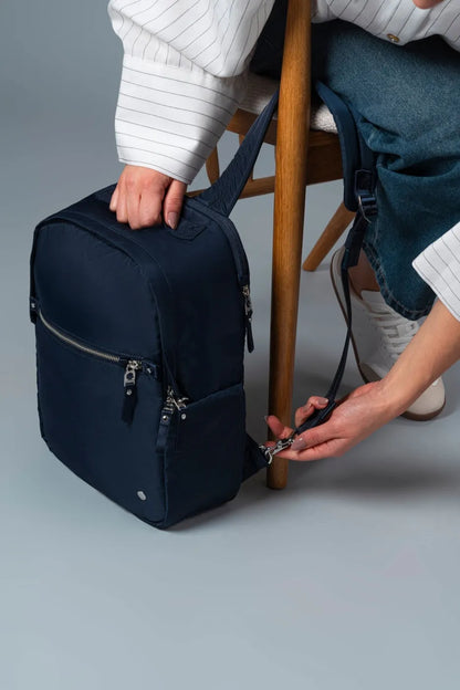 Pacsafe W 10L Anti-theft 13" Laptop Backpack in Navy, picked up by its top handle beside a chair, strap in the other hand