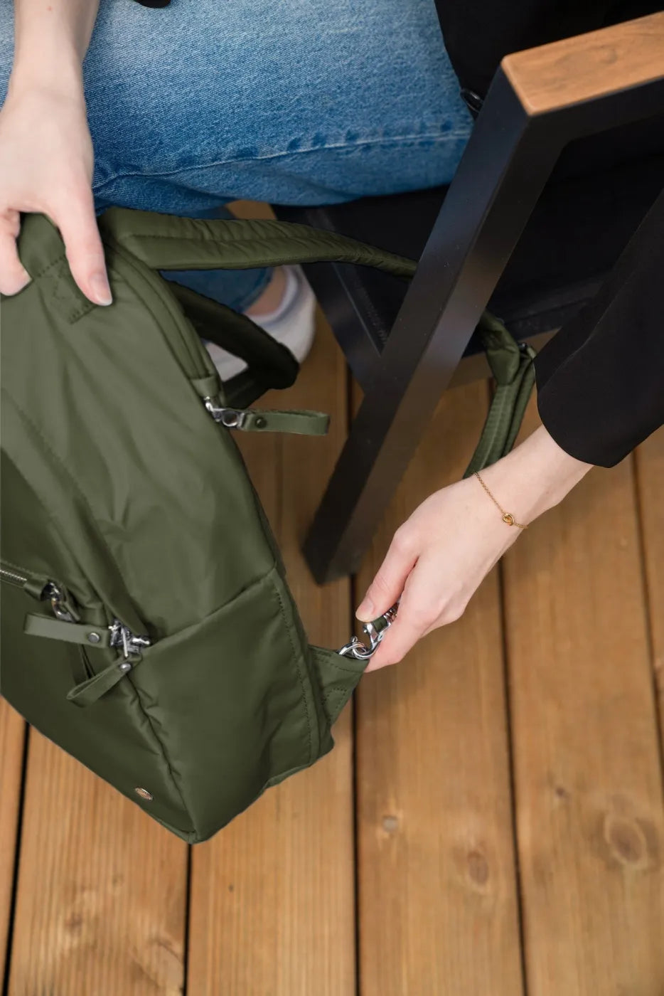 Pacsafe W 10L Anti-theft 13" Laptop Backpack in Olive, lifted by the strap from a timber deck beside a bench