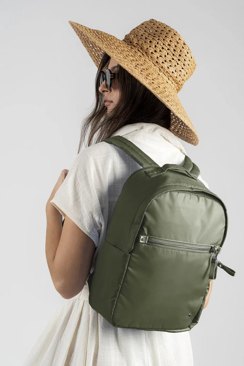 Pacsafe W 10L Anti-theft 13" Laptop Backpack in Olive, worn on one shoulder by a woman in a straw hat and white dress