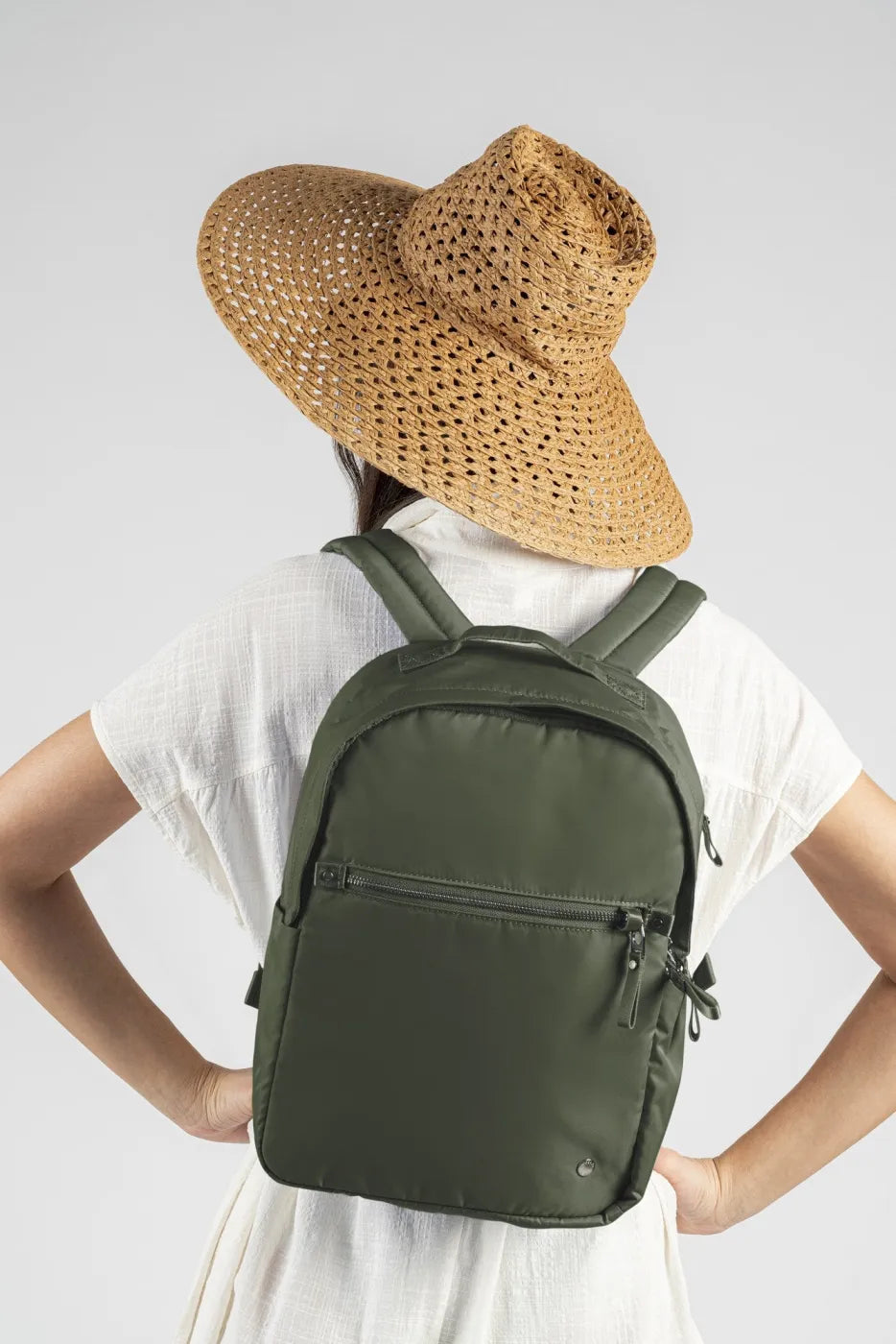 Pacsafe W 10L Anti-theft 13" Laptop Backpack in Olive, worn on both shoulders by a woman in a straw sun hat and white dress, seen from behind