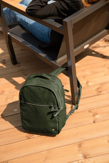 Pacsafe W 10L Anti-theft 13" Laptop Backpack in Olive, standing on a sunlit timber deck beside a bench