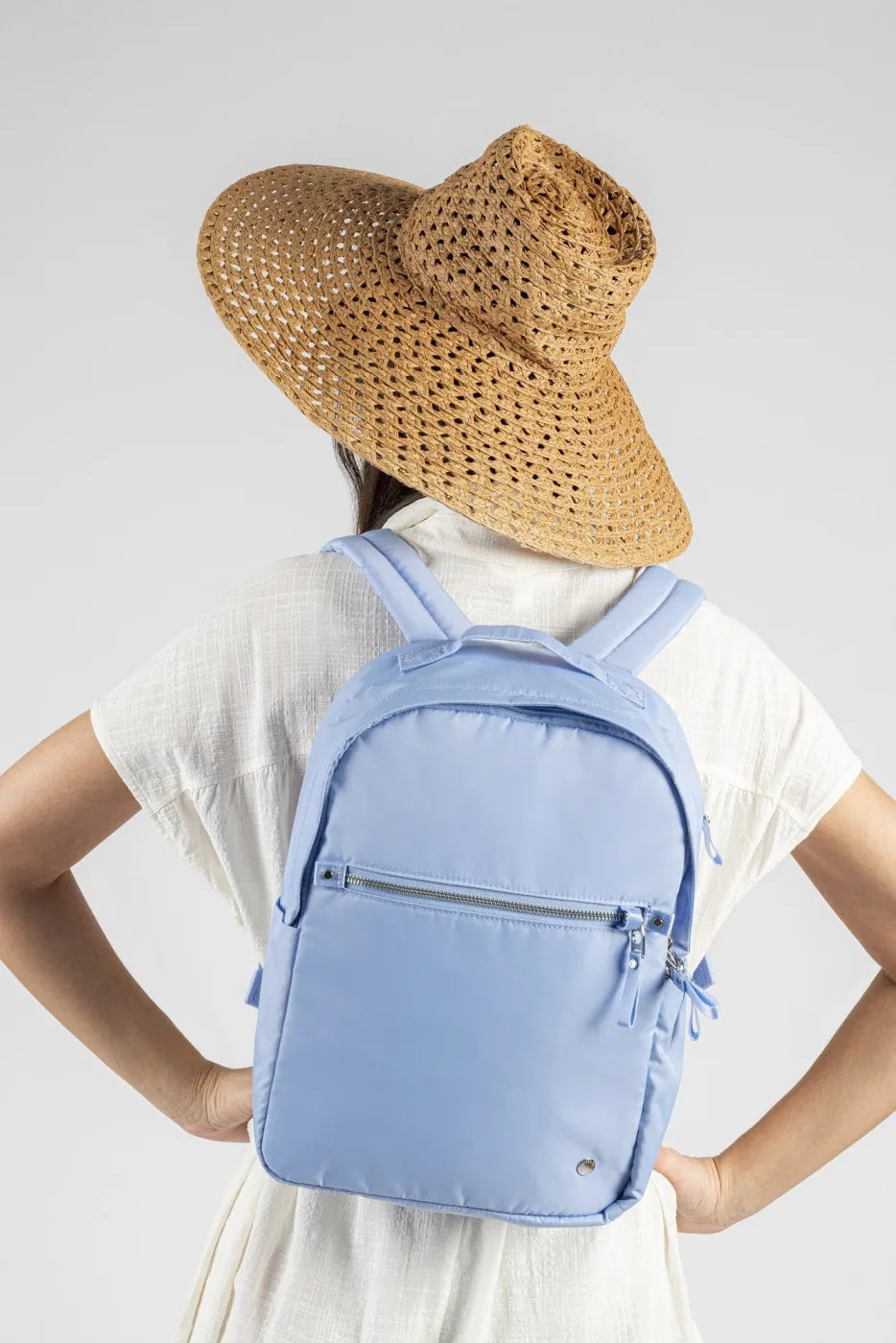 Pacsafe W 10L Anti-theft 13" Laptop Backpack in Serenity Blue, worn on both shoulders by a woman in a straw sun hat, seen from behind