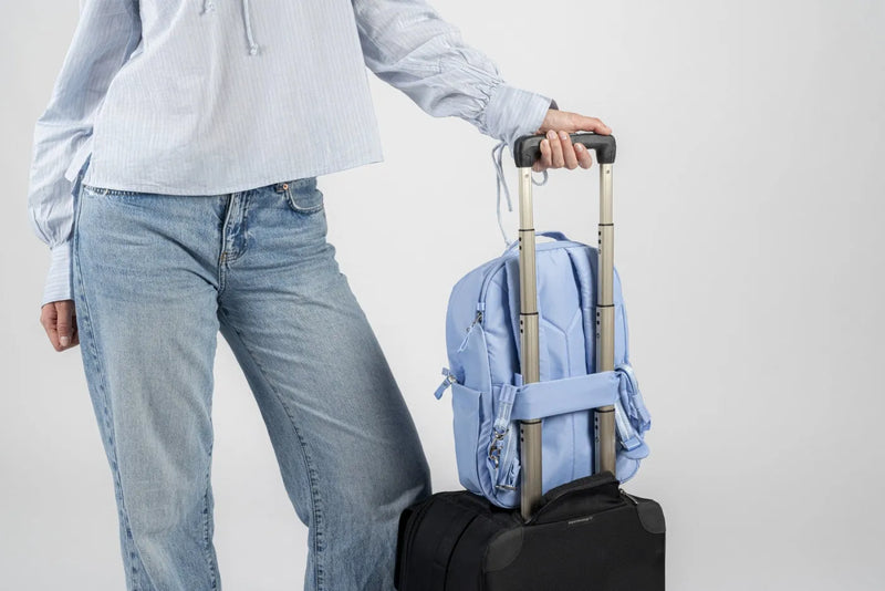 Pacsafe W 10L Anti-theft 13" Laptop Backpack in Serenity Blue, slid over the handle of a cabin case beside a woman in jeans