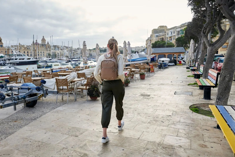 Pacsafe W 10L Anti-theft 13" Laptop Backpack in Taupe, worn on both shoulders by a woman walking a harbour promenade