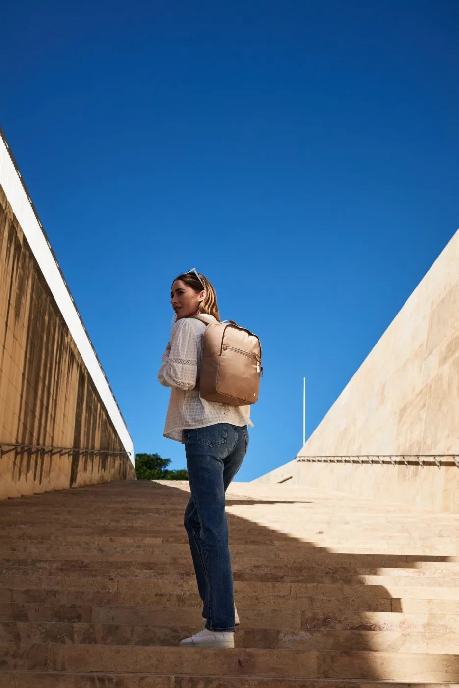 Pacsafe W 10L Anti-theft 13" Laptop Backpack in Taupe, worn on both shoulders by a woman climbing sunlit stone steps