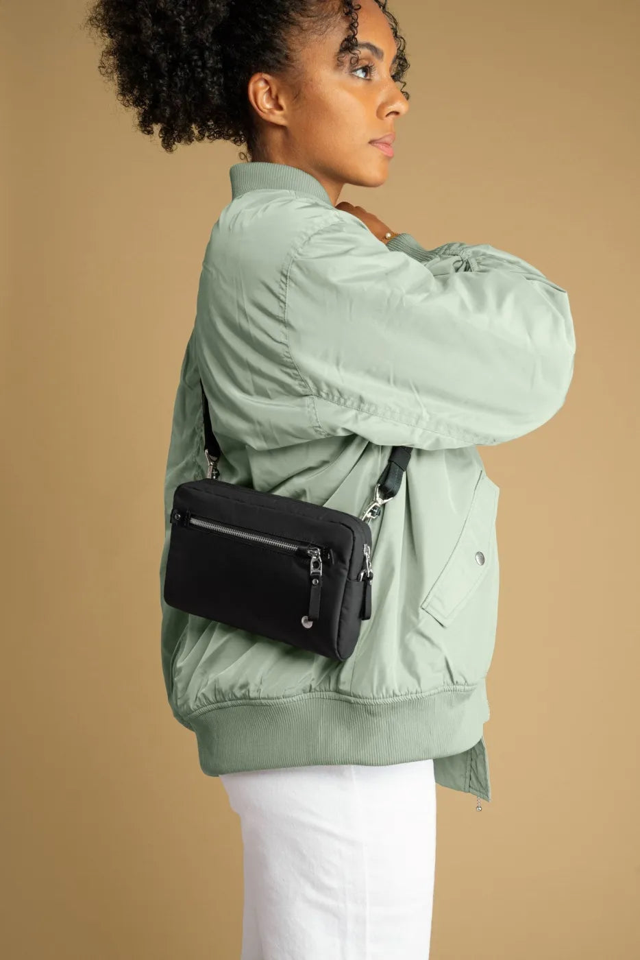 Pacsafe W Anti-theft 3-in-1 Sling Bag in Black, worn crossbody at the hip over a green bomber jacket, seen from the side