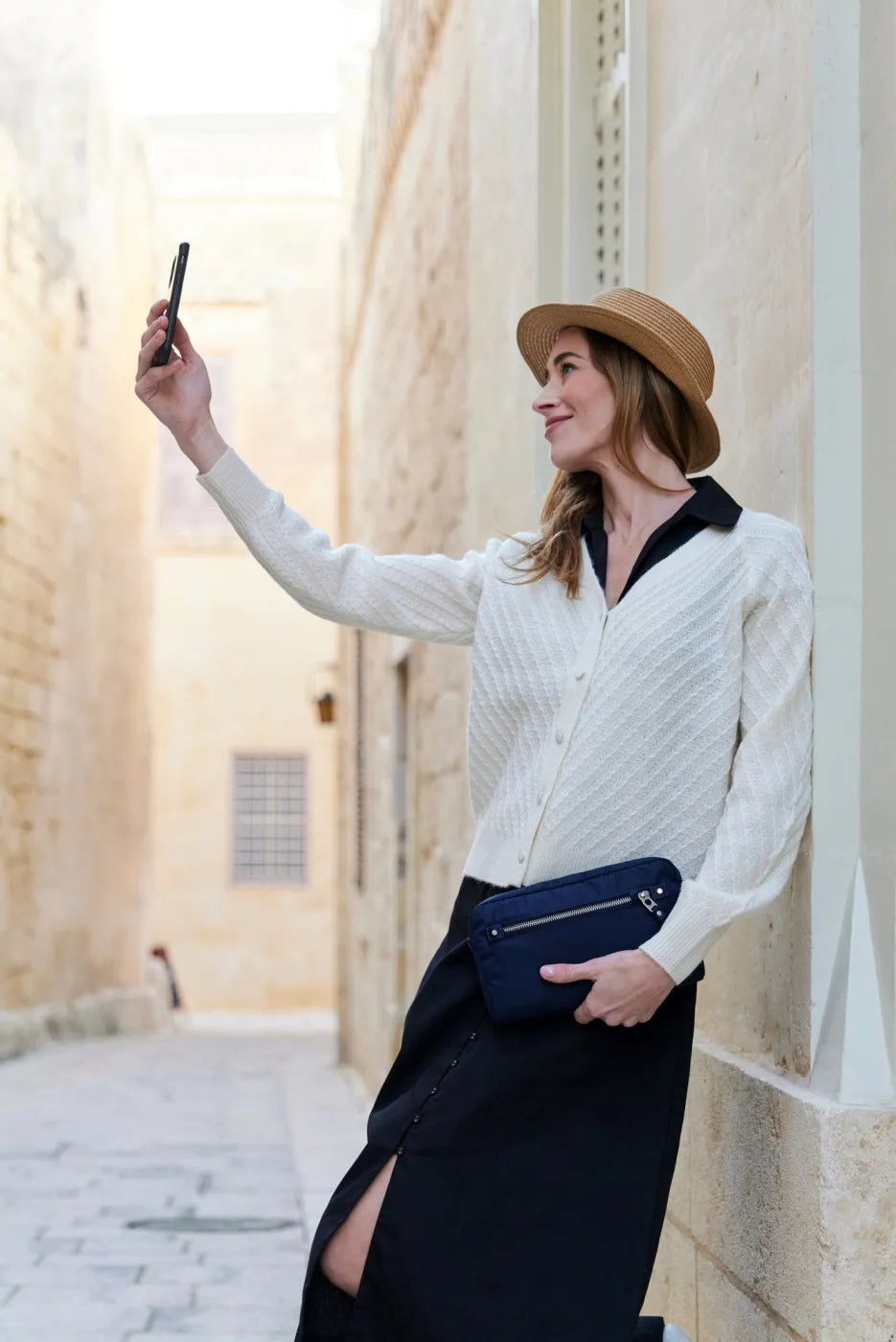 Pacsafe W Anti-theft 3-in-1 Sling Bag in Navy, worn at the waist by a woman taking a photograph in a stone alley
