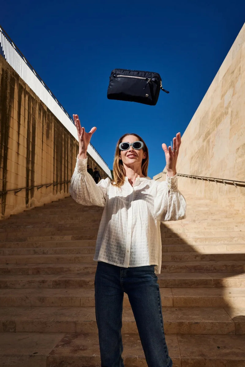 Pacsafe W Anti-theft 3-in-1 Sling Bag in Navy, tossed in the air by a woman standing on sunlit stone steps