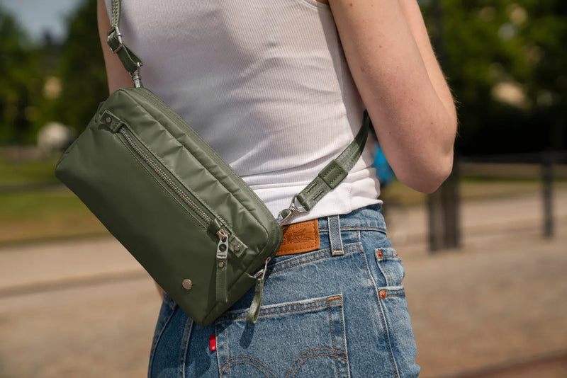 Pacsafe W Anti-theft 3-in-1 Sling Bag in Olive, worn at the back of the waist over jeans and a white tank, on a city street