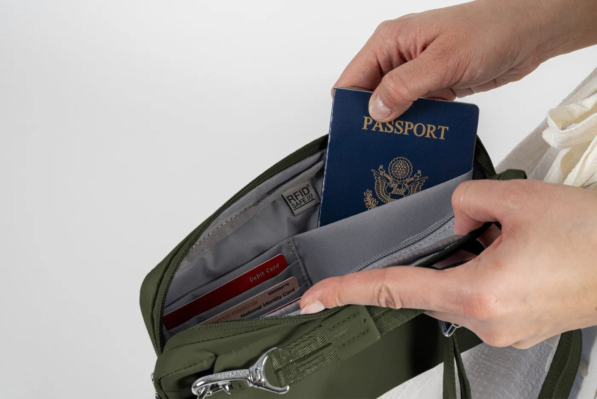 Pacsafe W Anti-theft 3-in-1 Sling Bag in Olive, held open on a studio backdrop, a passport being slid into the RFID pocket