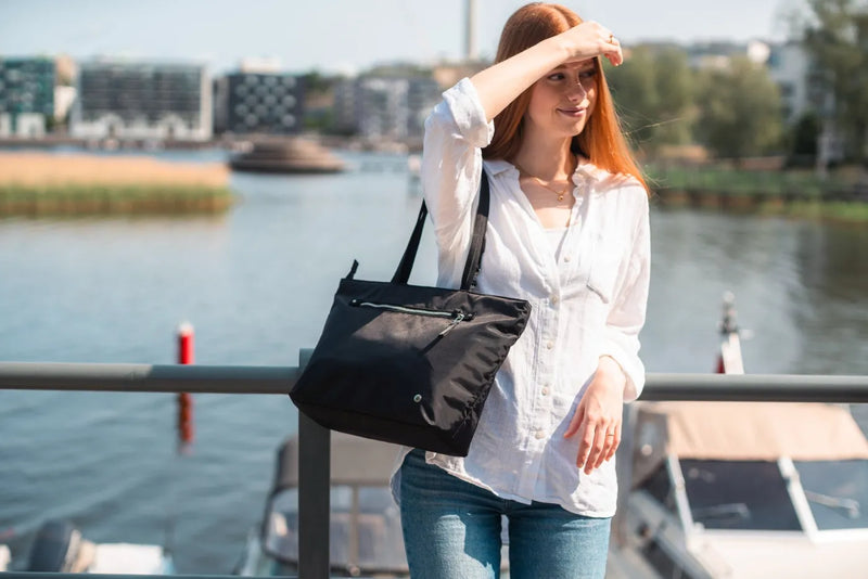 Pacsafe W Anti-theft Carryall 13" Laptop Tote in Black, carried on the shoulder by a woman standing at a waterfront railing