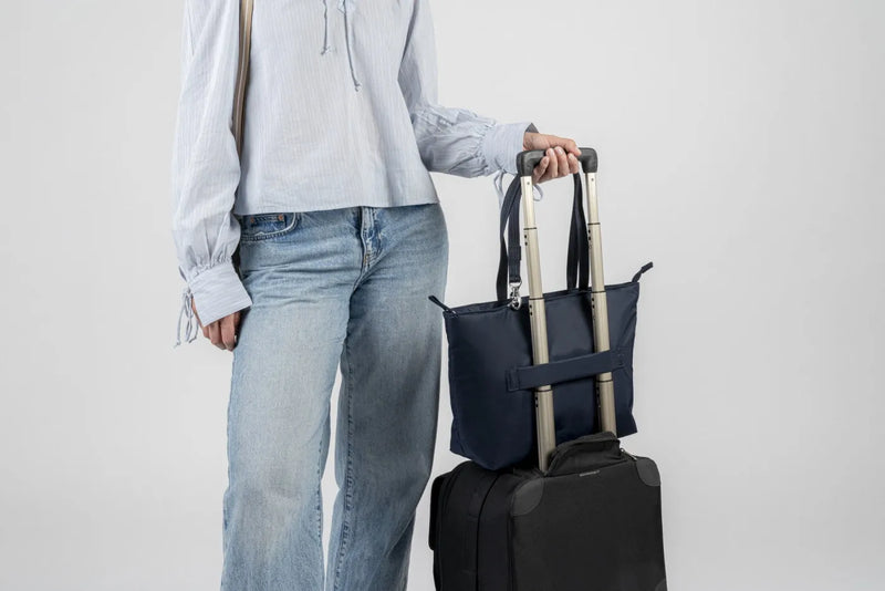 Pacsafe W Anti-theft Carryall 13" Laptop Tote in Navy, hooked over the handle of a cabin case beside a woman in jeans