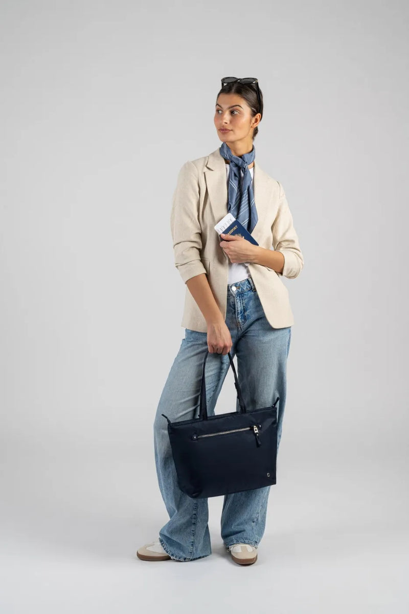 Pacsafe W Anti-theft Carryall 13" Laptop Tote in Navy, carried by its handles at the side of a woman holding a passport