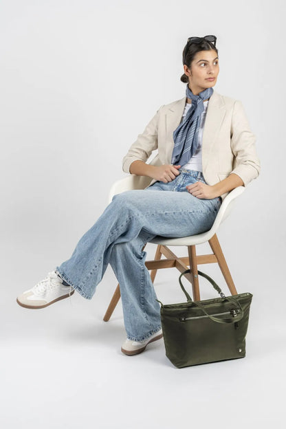 Pacsafe W Anti-theft Carryall 13" Laptop Tote in Olive, standing on the floor beside a woman seated in a cream blazer and jeans