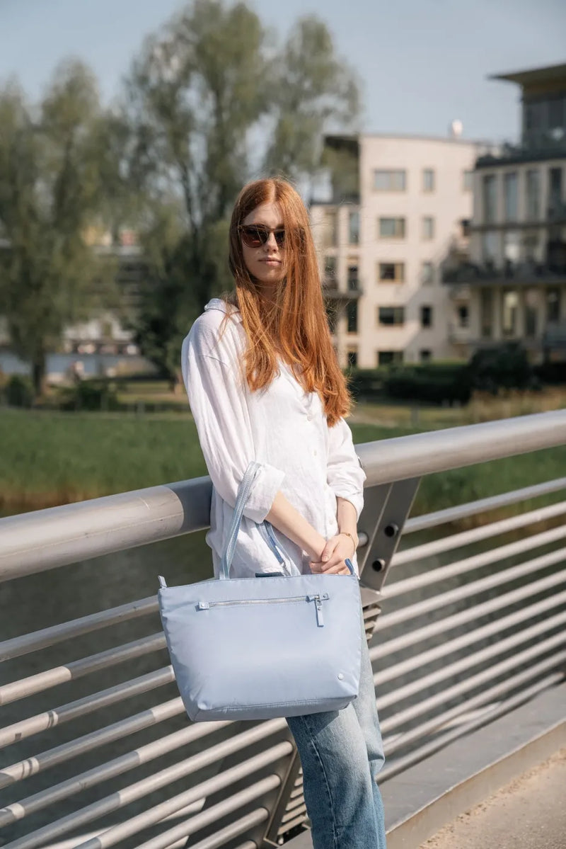 Pacsafe W Anti-theft Carryall 13" Laptop Tote in Serenity Blue, carried by a woman leaning on a bridge railing by the water