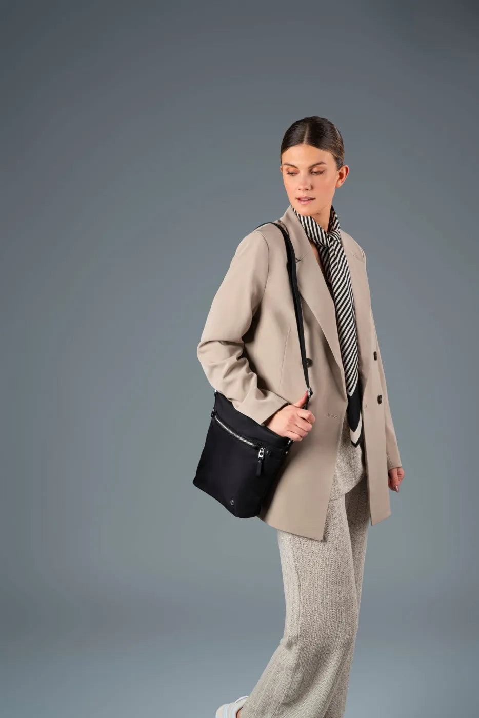 Pacsafe W Anti-theft Crossbody Bag in Black, worn crossbody under a beige blazer, hand on the strap