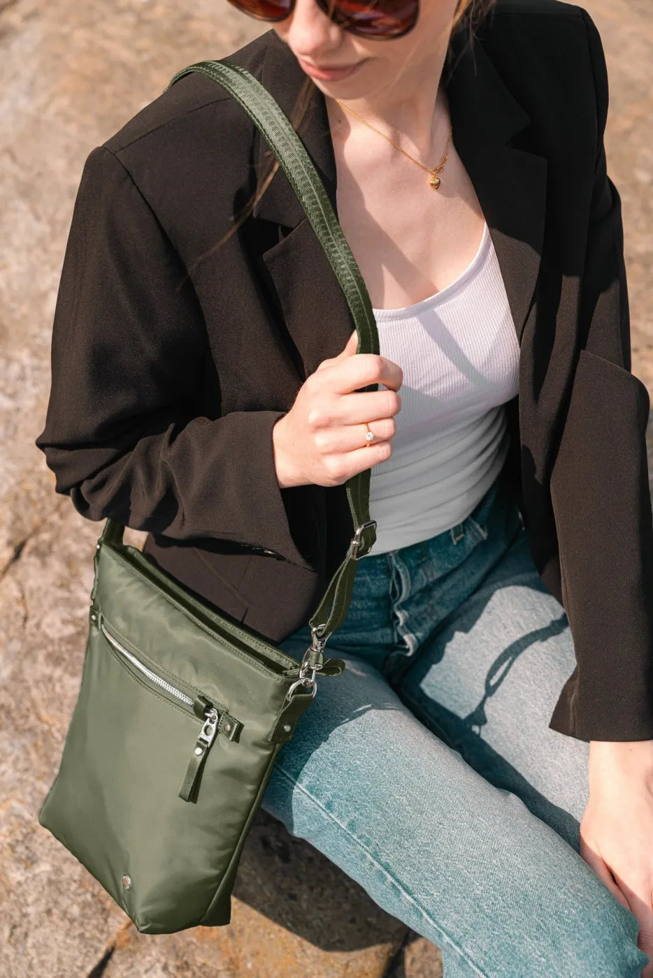 Pacsafe W Anti-theft Crossbody Bag in Olive, worn across the body by a woman in a black blazer and jeans, sitting on a rock