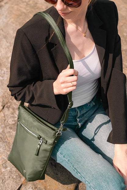 Pacsafe W Anti-theft Crossbody Bag in Olive, worn across the body by a woman in a black blazer and jeans, sitting on a rock
