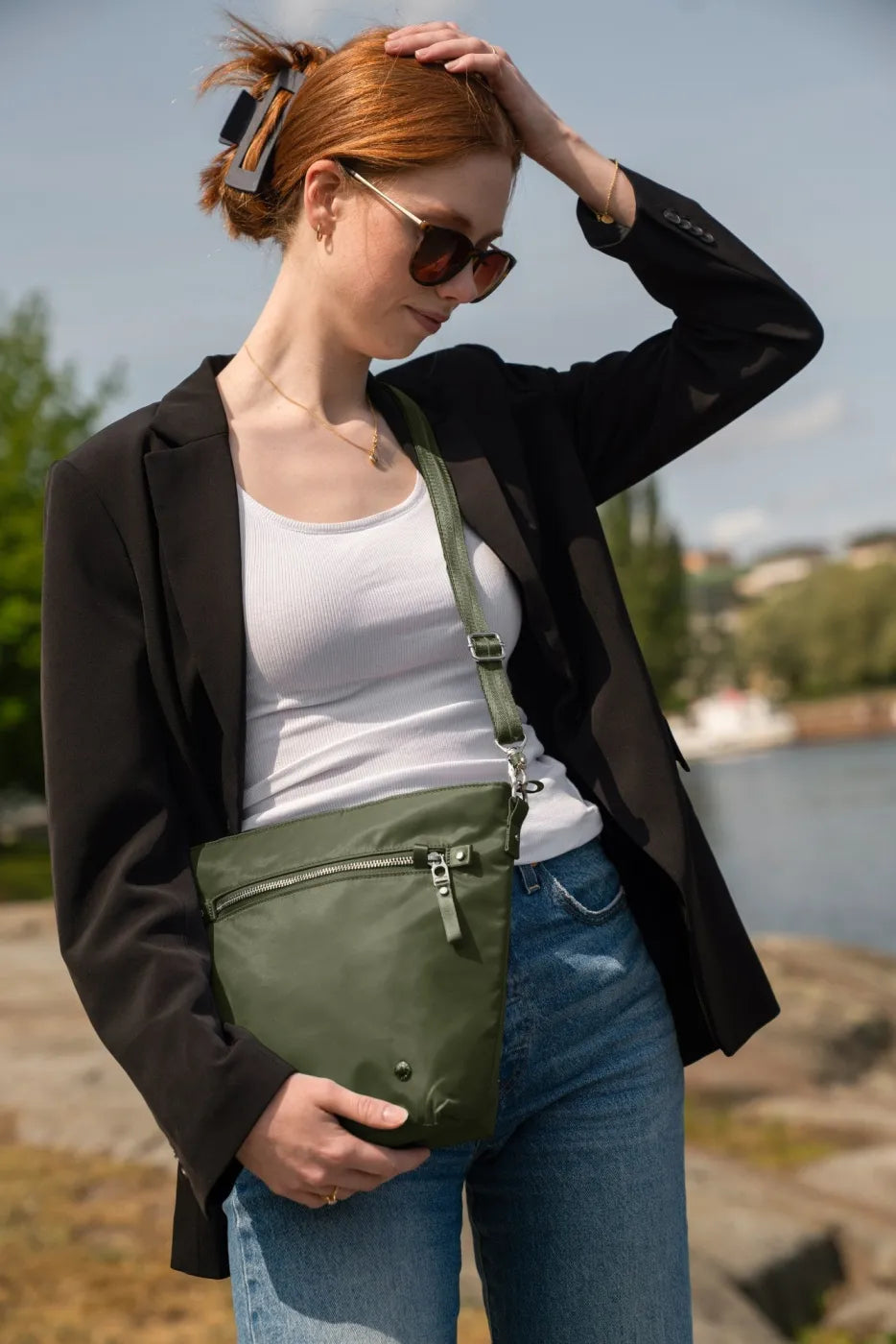Pacsafe W Anti-theft Crossbody Bag in Olive, resting at the hip of a woman in a black blazer and white tank by the water