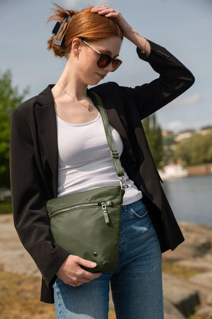 Pacsafe W Anti-theft Crossbody Bag in Olive, resting at the hip of a woman in a black blazer and white tank by the water