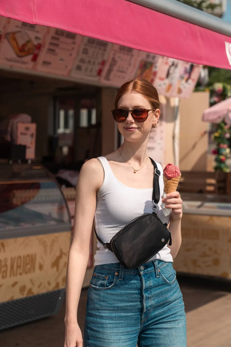 Pacsafe W Anti-theft Micro Sling in Black, worn crossbody by a woman with an ice cream outside an ice cream kiosk