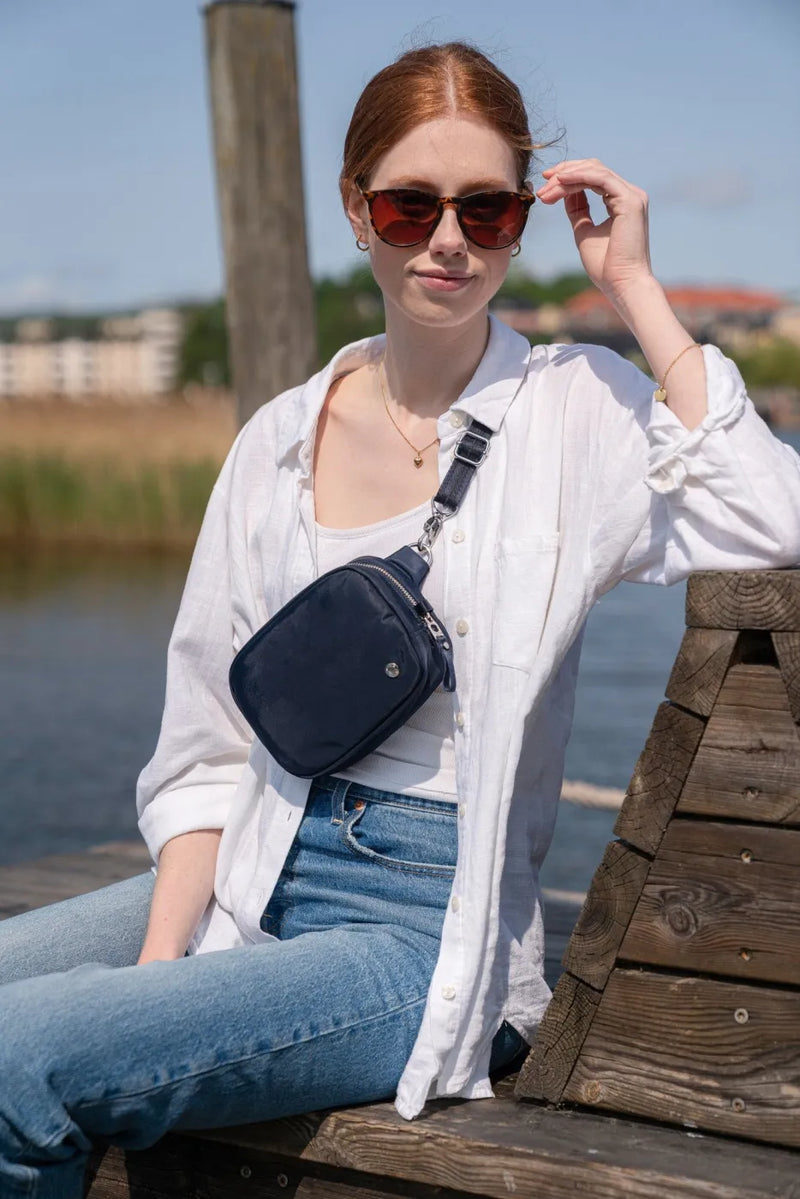 Pacsafe W Anti-theft Micro Sling in Navy, worn crossbody by a woman sitting on a jetty by the water