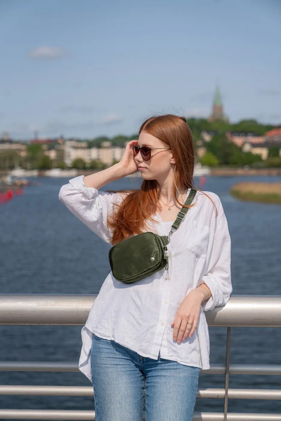 Pacsafe W Anti-theft Micro Sling in Olive, worn crossbody by a woman in a white linen shirt and jeans at a city waterfront
