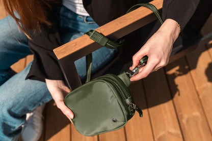 Pacsafe W Anti-theft Micro Sling in Olive, clipped onto its strap by a woman sitting on a boardwalk bench