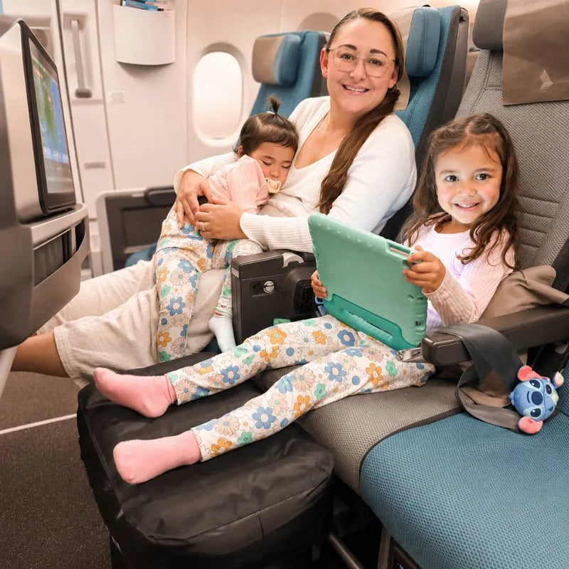 A woman and two children travel in a plane seat, with the woman holding a child while the girl in front holds a tablet with her legs resting on a Plane Pal inflatable travel cushion. The woman wears matching floral pajamas, and a blue plush toy rests on the seat beside them.