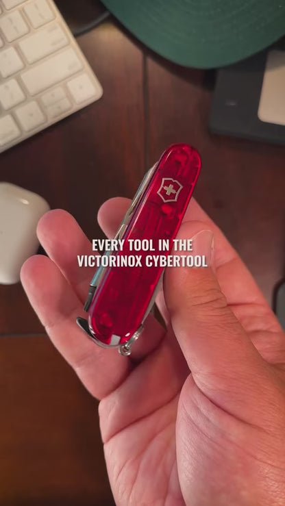 Victorinox CyberTool in translucent red on a wooden desk, opening the hex drive with its bit case, large and small blades, bottle opener and the pressurised ballpoint pen