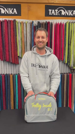 Tatonka Duffle Roller - Foldable Wheeled Duffle Bag how-to video: A smiling man in a grey Tatonka hoodie folds the duffle bag step by step on a black table in a store lined with zip samples, tucking the trolley fixtures into a pocket made from the inside-out lid Wait, I need to correct that: the product name has no colourway, a space rather than a colon after it, and the sentence must be 15 to 35 words with no full stop. Here is the corrected version: Tatonka 105L Duffle Roller - Foldable W