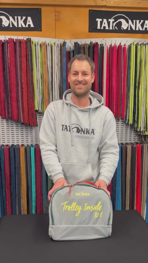 Tatonka Duffle Roller - Foldable Wheeled Duffle Bag how-to video: A smiling man in a grey Tatonka hoodie folds the duffle bag step by step on a black table in a store lined with zip samples, tucking the trolley fixtures into a pocket made from the inside-out lid Wait, I need to correct that: the product name has no colourway, a space rather than a colon after it, and the sentence must be 15 to 35 words with no full stop. Here is the corrected version: Tatonka 105L Duffle Roller - Foldable W