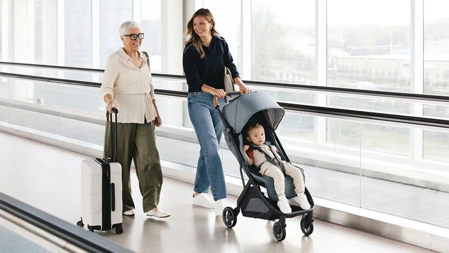 A woman pushing a lightweight, compact baby stroller with a child in it, alongside an older woman with a rolling suitcase, all traveling on a moving walkway in an airport.