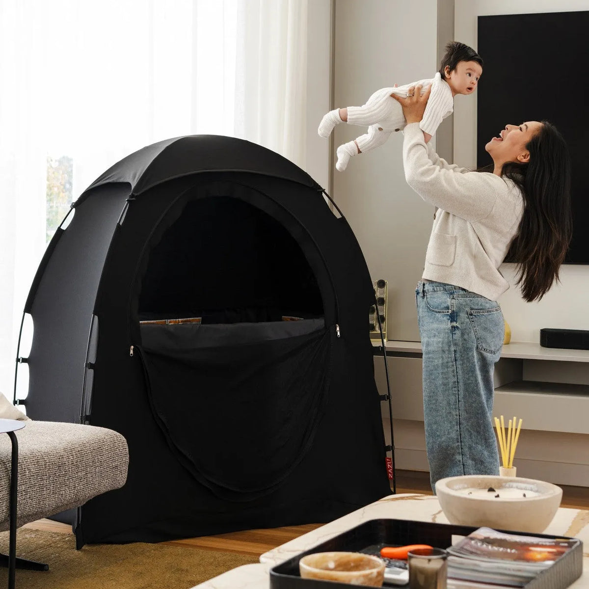 A woman in a living room holds a child above her head next to a black Zazu Rest Nest sleep tent. The room has a marble coffee table in the foreground, a gray sofa, and a large black television on the wall.