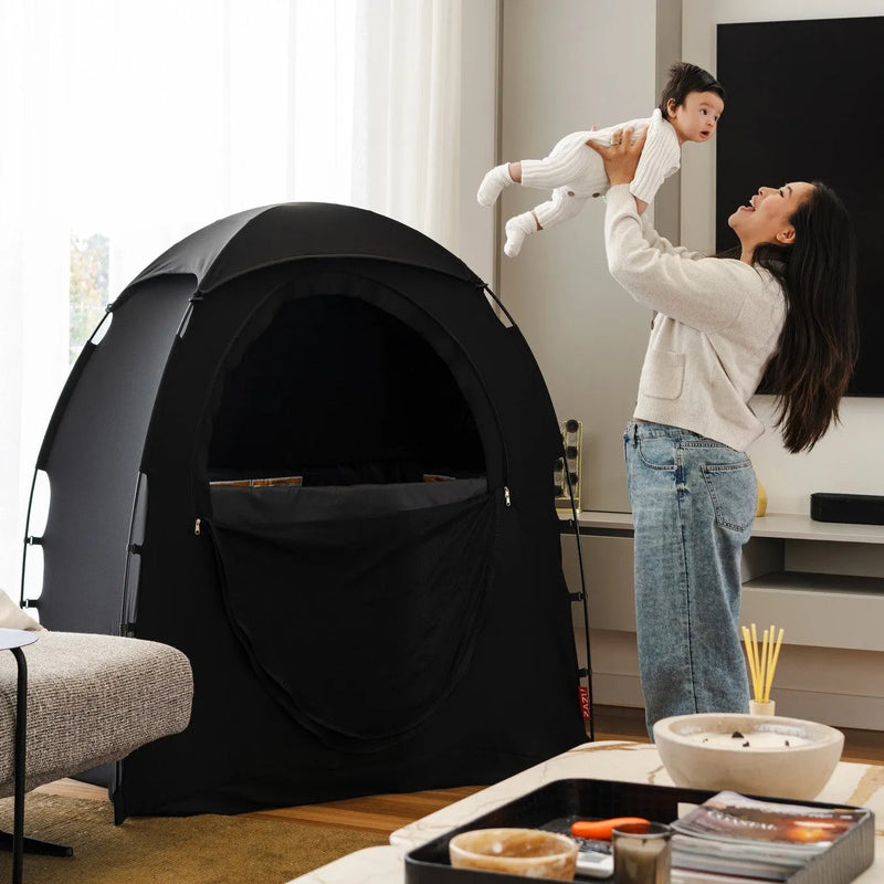 A woman in a living room holds a child above her head next to a black Zazu Rest Nest sleep tent. The room has a marble coffee table in the foreground, a gray sofa, and a large black television on the wall.